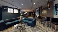 Located at 14909 Health Center Drive in Bowie, Maryland, this single-floor amenity space offers a sophisticated and well-appointed environment designed for socializing and recreation. The layout flows seamlessly through several distinct yet interconnected areas, unified by patterned striped carpeting and a consistent modern design palette of deep teal, charcoal, and warm neutral tones.

The main lounge area serves as the centerpiece of the space, featuring a sleek wall-mounted flat-screen TV above a linear gas fireplace framed in dark wood. Plush navy sectional seating and a glass-top coffee table create an inviting gathering spot, while a stylish multi-globe pendant light adds a contemporary focal point overhead. Large windows allow natural light to filter in, enhancing the open ambiance.

Adjacent to the lounge, a spacious billiards room provides a full-size pool table beneath an industrial-style pendant light fixture. A built-in bar counter with high stools runs along one wall, backed by a striking geometric teal tile accent wall and a wall-mounted television — ideal for entertaining.

The corridor connecting the areas features warm sconce lighting, tall decorative planters, and glass-panel accents, maintaining a polished, upscale feel throughout. High-end finishes, recessed lighting, and curated décor contribute to an atmosphere that is both functional and refined.