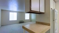 This single-floor space integrates a kitchen and open living area into one cohesive layout. The kitchen area features warm wood-toned countertops and upper cabinetry with dark trim, providing functional storage and prep space. The living area is finished with neutral carpet and light green walls, creating a calm, understated atmosphere. Two windows along the far wall allow natural light to fill the room, lending an airy quality to the interior. A wall-mounted air conditioning unit ensures climate control year-round. The textured ceiling adds a subtle design element consistent with the space's practical, straightforward aesthetic. Overall, the space offers a simple, functional environment well-suited for everyday living.