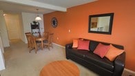 This single-floor space makes a strong visual impression with its bold orange accent wall, which serves as the focal point of the open-concept living and dining area. The living room is comfortably appointed with a dark leather sofa accented by warm-toned throw pillows that complement the striking wall color. A round wooden coffee table anchors the seating area, while plush carpeting adds warmth underfoot throughout the space.

A large framed mirror on the accent wall enhances the sense of depth and reflects light into the room, offering a glimpse of an adjacent kitchen area. The open layout flows naturally into the dining area, where a round wooden dining table is surrounded by matching wood chairs, creating an inviting space for meals and gatherings. A pendant light fixture hangs above the dining table, providing focused illumination and an element of understated style.

A wall-mounted clock and a shelving unit with decorative accents add personality to the space. The overall ambiance is warm and energetic, with the orange tones creating a lively yet cohesive aesthetic throughout the combined living and dining area.