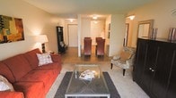 This single-floor residential unit offers a thoughtfully designed open-concept layout that connects the living, dining, and kitchen areas in a seamless and functional arrangement. The living area features warm, neutral-toned walls complemented by light carpet underfoot, creating a cozy and inviting atmosphere. A glass-top coffee table sits atop a charcoal area rug, anchoring the seating space that includes a rich red sofa and an eye-catching zebra-print accent chair, adding a touch of personality to the room. Hardwood-style flooring transitions into the dining area, where a set of elegant dark wood chairs surrounds a dining table positioned beneath flush-mount ceiling lighting. The kitchen is visible beyond the dining space, separated by a partial wall that maintains the open feel while defining each zone. A hallway extending to the right suggests additional private spaces such as bedrooms and a bathroom. Near the entry, a dark wood bookcase adds storage and character. The space is well-lit throughout with a combination of ambient ceiling fixtures and a classic table lamp, contributing to a warm, comfortable ambiance suitable for everyday living.