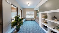 Located at 6822 Shannon Pky in Union City, Georgia, this single-floor living room offers a carefully curated lounge environment designed for comfort and community. The space is defined by plush, patterned carpeting in cool blue-grey tones that anchors the room with a sense of warmth and style. A built-in fireplace with a marble surround serves as a natural focal point, flanked by white open shelving that provides both decorative and functional storage. On the opposite side, a cozy built-in bench with striped cushions sits beneath a window overlooking lush greenery, inviting natural light into the interior. Stylish wingback chairs with tweed upholstery and accent pillows are thoughtfully arranged throughout, offering comfortable seating throughout the space. Interior windows with dark bronze frames provide views into an adjacent common area featuring a striking coffered ceiling, enhancing the sense of openness and connectivity. Flush-mount pendant lighting illuminates the space with a warm glow, while accent walls in sage green and taupe contribute to a calming, sophisticated palette. Lush indoor plants add a refreshing natural touch. This living room strikes a harmonious balance between elegance and everyday comfort.