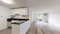 Located at 950 E Center St in Wallingford, Connecticut, this single-floor residence offers a clean, modern aesthetic throughout. The open-concept layout seamlessly connects the kitchen and living area, creating a cohesive and functional living space. Light-toned hardwood-style flooring runs throughout the main living areas, lending warmth and a sense of continuity.

The kitchen is thoughtfully designed with crisp white cabinetry, dark countertops, and stainless steel appliances, including a range, dishwasher, and refrigerator. Tile flooring anchors the kitchen area, providing a practical contrast to the wood floors of the adjoining spaces.

The spacious living area is bathed in natural light, courtesy of a large sliding glass door that opens to a private outdoor patio area surrounded by lush greenery. A wall-mounted AC unit and baseboard heating ensure year-round comfort. The bedroom also features a sliding glass door to the outdoors, offering additional natural light and direct exterior access beneath a covered patio structure.

The bathroom is finished with neutral tile flooring and includes a full tub-shower combination, toilet, and a vanity with storage. Built-in closet storage in the bedroom adds to the unit's practicality. The overall color palette of whites and light neutrals creates a bright, airy ambiance throughout the entire space.