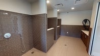 Located in Dallas, this single-floor facility is purpose-built for pet washing and grooming, offering a thoughtfully designed space that balances practicality with a clean, modern aesthetic. The interior is defined by richly toned dark brown subway tile that lines the walls to mid-height throughout, creating a durable and visually cohesive environment. Light-colored large-format floor tiles contrast the darker wall tile, contributing to a bright, hygienic feel underfoot.

Multiple open wash stations are arranged with low tiled partition walls, each equipped with chrome handheld shower fixtures and built-in ledges for storing grooming supplies. Floor drains are strategically positioned at each station to ensure efficient water management. Recessed lighting illuminates the space evenly throughout, while a ventilation system with ceiling-mounted HVAC vents maintains air quality and comfort.

The entry and transitional areas feature glass-paneled sliding doors that allow natural light to filter into the space from nearby windows, enhancing visibility and openness. A dispensing station is also present, providing convenient access to grooming products. Security cameras are installed throughout, ensuring a safe and monitored environment. Overall, the facility presents a clean, well-organized, and professional space designed for efficient and comfortable pet care.