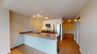 This well-designed single-floor unit offers a thoughtful layout that balances open communal spaces with private rooms, all unified by warm hardwood flooring throughout. The open-concept kitchen serves as a central hub, showcasing a large island with a gray stone countertop, an undermount sink, and stainless steel appliances including a refrigerator, range, and built-in microwave. Crisp white upper cabinetry extends to the ceiling, complemented by warm wood lower cabinets and a classic subway tile backsplash. Track lighting overhead adds both task and ambient illumination. The adjacent living area is generously proportioned, offering a bright and airy atmosphere that flows seamlessly from the kitchen. Floor-to-ceiling windows in the primary bedroom frame impressive city views, filling the space with abundant natural light. A secondary bedroom provides additional flexibility, accessed via a frosted-glass sliding door that enhances the modern aesthetic while maintaining privacy. The bathroom is clean and functional, featuring a wood-toned vanity with a stone countertop, undermount sink, full-width mirror, and a soaking tub with a shower. The neutral color palette and contemporary finishes throughout create a cohesive, modern living environment.