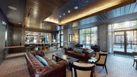 This single-floor lounge space masterfully combines industrial character with refined, classic elegance. The flooring throughout is laid in a distinctive herringbone brick pattern, grounding the space with warmth and texture. A richly stained wood-planked ceiling with recessed lighting and a softly illuminated tray detail crowns the main seating area, adding depth and a warm ambient glow.

The lounge is anchored by a full bar equipped with brass-accented high stools, backlit shelving displaying glassware, and a luminous countertop. Nearby, tufted brown leather Chesterfield sofas and mixed accent chairs define a generous seating area centered around dark coffee tables. A wall-mounted flat-screen television adds a modern touch to the otherwise vintage-inspired setting.

An ornate marble fireplace serves as a focal point along the exposed brick accent wall, flanked by a built-in wooden bookshelf and intimate café-style seating. Vintage signage, including a prominent Campbell & Co. Edinburgh Ales advertisement, adds layers of character. Floor-to-ceiling steel-framed windows flood the space with natural light and provide views of a verdant outdoor patio, while sheer curtains offer privacy without sacrificing luminosity. The overall ambiance is inviting, clubby, and distinctly curated.