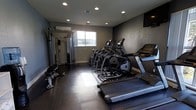 This fitness center is a dedicated, single-floor workout facility designed with functionality and comfort in mind. The space is finished with dark hardwood-style flooring that contrasts cleanly against the neutral gray walls, creating a focused and energizing atmosphere. Recessed ceiling lighting provides bright, even illumination throughout the room, ensuring a well-lit environment for all types of exercise.

The cardio section is well-stocked, featuring multiple treadmills, elliptical trainers, and a stair climber, all arranged to maximize the use of the available floor space. A wall-mounted television is positioned for convenient viewing during cardio sessions. On the strength training side, the facility includes a multi-functional cable and pulley machine, along with additional free-standing equipment to support a variety of resistance exercises.

A wall-mounted split air conditioning unit ensures the space remains climate-controlled year-round. Natural light filters in through a window along one wall, adding a refreshing element to the interior. A mirrored section near the strength equipment allows users to monitor their form during workouts. Overall, this fitness center offers a complete and practical environment suited for a wide range of fitness routines.