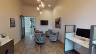 Located in Brighton 3050, this single-floor space functions as a well-designed business centre combining comfort with functionality. The interior is characterised by impressively high ceilings adorned with elegant crown moulding, warm neutral walls, and rich dark hardwood-style flooring that runs throughout, creating a cohesive and professional atmosphere.

The lounge area serves as a central gathering point, furnished with a cluster of patterned geometric armchairs arranged around small round side tables. A wall-mounted flat-screen television anchors one corner, while a striking large-format equestrian artwork adds a bold decorative focal point. Overhead, a cascading pendant light fixture with exposed globe bulbs provides warm, ambient illumination and contributes to the space's stylish character.

Adjacent to the lounge, a dedicated workstation area offers multiple private computer cubicles equipped with desktop computers, separated by frosted-panel dividers for focused individual work. A printing station is conveniently positioned nearby. Natural light fills this section through tall windows dressed with blinds, complemented by a café-style table and seating for informal collaboration.

Glass-panel doors connect the zones seamlessly, enhancing the open flow between areas. Overall, the space balances a welcoming, social atmosphere with practical, productivity-focused amenities.