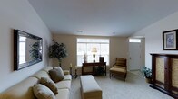 This single-floor residence offers a comfortable and well-appointed layout designed for easy living. The living room serves as the heart of the home, bathed in natural light streaming through a large picture window with a transom panel above, creating an airy and open atmosphere. Neutral-toned walls and plush wall-to-wall carpeting lend a warm, cohesive feel throughout the space. The room is thoughtfully arranged with a cushioned sofa and coordinating ottoman, complemented by a classic wooden accent table adorned with a traditional lamp and floral arrangement. A woven armchair with a matching footstool adds a cozy reading nook near the window, while a tall indoor plant brings a touch of greenery and life to the corner. Decorative candle holders provide an elegant accent. The entryway is visible from the living area, featuring a sidelight transom window above the front door that allows additional natural light to filter in, reinforcing the bright and welcoming character of the home. The overall ambiance is classic and serene, with a neutral palette and traditional design elements that create a timeless, comfortable living environment.