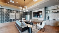 Located at 9600 W Georgia Ave in Glendale, Arizona, this single-floor community space offers a thoughtfully designed interior that blends modern aesthetics with comfortable functionality. The combined living and dining area is generously proportioned, providing ample room for both relaxation and social gatherings.

The living area is a standout feature, showcasing a coffered wood-paneled ceiling with a striking multi-globe chandelier that anchors the space. A large wall-mounted flat-screen TV is framed by a light stone surround, complemented by built-in wood cabinetry below. A bold, textured dark accent wall adds depth and visual interest, while warm wood-plank flooring runs throughout. Seating is arranged around a layered area rug with a mix of plush sofas, accent chairs, and sculptural side tables, creating an inviting lounge atmosphere.

The dining area transitions seamlessly from the living space, featuring a rustic wood communal table paired with mixed industrial-style chairs. Large windows along the dark-painted walls allow natural light to filter in, offering views of the surrounding exterior. A glass-paneled door provides direct access to the outdoor courtyard area.

Throughout the space, curated wall art, open shelving, and refined architectural details contribute to a cohesive, upscale ambiance suited for both casual gatherings and structured events.