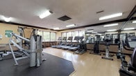This fitness center offers a comprehensive workout environment designed to accommodate a range of exercise routines. The space is laid out on a single level, with light wood-tone flooring throughout and rubberized mat sections strategically placed beneath the strength training equipment to provide cushioning and protection.

The cardio zone is a standout feature, housing a row of treadmills and at least one stair climber positioned along a bank of large windows. These windows frame views of mature trees outside, providing a pleasant natural backdrop and ample natural light during daytime use. The placement of the cardio machines directly in front of the windows creates an open, airy feel that enhances the workout experience.

On the opposite side of the room, a full-length wall mirror visually expands the space and allows users to monitor their form. The strength training area includes a multi-station cable machine, a weight bench with a press attachment, and additional resistance equipment, offering options for both upper and lower body training.

Overhead fluorescent lighting ensures the room is well-lit at all hours. The textured ceiling and dark wood trim detailing along the upper walls add a classic finishing touch to the overall design of the space.