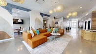 This thoughtfully designed amenity floor spans a generous footprint, offering residents a diverse range of communal spaces united by a cohesive contemporary aesthetic. The centerpiece is a large open lounge area featuring polished concrete floors, soaring ceilings, and striking globe pendant lights with woven, honeycomb-patterned shades that cast a warm, inviting glow. A bold abstract mural adorns the walls, complementing the rich caramel leather sectional sofa accented with teal and gold throw pillows. A wood-paneled media wall with a mounted flat-screen television adds warmth and texture to the seating area, while wicker accent chairs provide additional casual seating throughout.

A dedicated dining and coworking nook offers bar-height seating and a wall-mounted screen, ideal for both casual meals and productive work sessions. Glass-enclosed conference rooms provide private meeting spaces, furnished with round tables and upholstered chairs.

A game area features a shuffleboard table beneath an artistic industrial-style pendant light installation, adding a playful touch. The hallway connecting these spaces is lined with polished floors and decorative artwork, leading to dual elevators framed by warm wood-paneled walls.

The fitness center is well-appointed with cardio machines including treadmills, ellipticals, a rowing machine, and a stationary bike, alongside a comprehensive strength training station and a full dumbbell rack. Floor-to-ceiling mirrors and a large garage-style window overlooking the outdoors complete the bright, energizing workout environment.