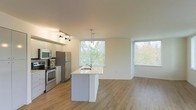 This single-floor space showcases an open-concept layout that seamlessly connects the kitchen and living area, creating a spacious and airy atmosphere. The kitchen is well-appointed with stainless steel appliances, including a refrigerator, electric range, and over-the-range microwave, all set against crisp white cabinetry. A central island with a light stone countertop and an integrated sink serves as both a functional workspace and a natural dividing point between the kitchen and living areas. A sleek pendant light hangs above the island, complementing the track lighting positioned over the kitchen. The living area is bathed in abundant natural light, courtesy of two generously sized windows that frame views of surrounding greenery and trees, enhancing the connection to the outdoors. Light wood-tone flooring runs throughout the space, adding warmth and continuity. The neutral white walls and clean lines contribute to a modern, minimalist aesthetic. Overall, the space offers a welcoming and functional environment with a bright, contemporary feel.