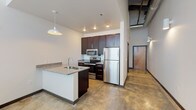 This studio flat blends modern finishes with industrial-loft aesthetics across a single open-plan level. Polished concrete floors run throughout, anchoring the space with a raw, contemporary feel that complements the exposed brick accent wall found in the main living area. Large windows fitted with horizontal blinds allow natural light to fill the room, while a ceiling fan overhead adds comfort and airflow.

The kitchen is well-appointed and centrally positioned, featuring dark espresso cabinetry, granite-look countertops, and a full suite of stainless steel appliances including a refrigerator, electric range, and built-in microwave. A peninsula with an integrated sink and additional cabinetry below extends the workspace and provides a natural division within the open layout. Industrial-style pendant lighting overhead adds character and focused illumination.

The bathroom is clean and functional, outfitted with a vanity featuring granite-look countertops and dark cabinetry, a full tub/shower combination, and a toilet — all set against bright white walls. A dedicated utility area houses a stacked washer and dryer unit alongside open wire shelving, offering practical in-unit laundry convenience. Exposed ductwork visible in portions of the space further reinforces the industrial design language throughout.
