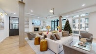 Located at 17-00 Pollitt Dr in Fair Lawn, NJ, this thoughtfully designed single-floor space offers a seamless open-plan layout combining a spacious living room and a well-appointed kitchen. The living area is anchored by a striking stone-surround fireplace topped with a wall-mounted flat-screen TV and flanked by built-in floating shelves, creating a refined yet functional focal point. Light wide-plank hardwood flooring flows throughout, complementing the clean white walls and lending a warm, airy atmosphere to the entire space. Floor-to-ceiling black-framed glass doors and expansive windows flood the interior with natural light while providing clear views of the surrounding exterior. Structural columns with warm wood accents add architectural character, while a stylish multi-arm brass pendant light anchors the seating area below. The kitchen features sleek, dark wood cabinetry paired with crisp white marble-look countertops, an undermount stainless steel sink with a matte black faucet, and framed mirror-front upper cabinets that enhance the sense of depth. Recessed lighting throughout ensures consistent illumination across both areas. The overall ambiance is modern and sophisticated, balancing neutral tones with warm metallic accents for an inviting and polished environment.