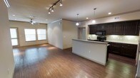 This single-floor residence offers a thoughtfully designed open-concept layout that effortlessly connects the kitchen and living areas into one cohesive, flowing space. Rich hardwood flooring extends throughout, lending warmth and a sense of continuity to the interior.

The kitchen is a standout feature, anchored by dark espresso cabinetry that contrasts elegantly with a light granite or stone countertop peninsula. The peninsula doubles as a breakfast bar, providing additional seating and workspace. Black appliances, including a range and microwave, complement the cabinetry, while pendant lighting suspended above the peninsula adds both task illumination and a stylish focal point.

The adjacent living area is generously proportioned, offering ample room for a variety of furniture arrangements. A ceiling fan with light fixture enhances comfort and air circulation. Track lighting along the ceiling provides additional ambient and accent illumination. Recessed lighting throughout the space ensures even, well-distributed light.

Natural light filters in through windows fitted with horizontal blinds, brightening the living area. Crown molding details add a refined architectural touch to the ceilings. The overall ambiance is modern yet inviting, with a neutral color palette that provides a versatile backdrop for any interior style.
