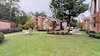 This single-story shared laundry facility is situated within a multi-building residential community characterized by classic brick exteriors, tan siding accents, and lush, well-maintained grounds. Mature trees and neatly trimmed hedgerows frame winding walkways that connect the buildings, creating a welcoming and park-like outdoor atmosphere.

The laundry facility itself is accessed through a glass-paneled entry door that allows natural light to filter into the space. The interior features light wood-look flooring throughout, neutral light gray walls, and crown molding details that contribute a clean, finished appearance. A ceiling fan with integrated lighting provides ventilation and illumination across the room.

The facility is well-equipped with multiple commercial-grade top-loading washing machines by Speed Queen arranged along one wall, complemented by a row of stacked dryer units lining the opposite side. Informational signage is posted throughout for user guidance. A wall-mounted air conditioning unit ensures comfort during warmer months.

Large windows along the walls allow views of the surrounding greenery and neighboring buildings, maintaining a bright and airy feel within the space. The overall layout is functional and spacious, offering residents convenient and comfortable access to laundry amenities in a tidy, well-kept environment.