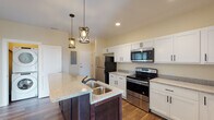 Located at 1900–1914 Atchison Ave in Hammond, Indiana, this single-floor unit offers a thoughtfully designed interior with modern finishes throughout. The kitchen serves as a central highlight, anchored by a large dark-base island with granite countertops and a double-basin sink, illuminated by two industrial-style pendant lights. White shaker-style cabinetry lines the walls, providing ample storage, while stainless steel appliances—including a refrigerator, electric range, and over-the-range microwave—complete the functional and stylish cooking space. Rich hardwood-style flooring flows throughout the main living areas, adding warmth and continuity to the open layout. The living area is bright and inviting, benefiting from natural light and views of the surrounding neighborhood through large windows. The floor plan also includes well-sized bedrooms with hardwood-style flooring and closet access. Two bathrooms serve the unit: one featuring a walk-in shower with a granite vanity top and large mirror, and another with a full soaking tub and shower combo. Warm neutral wall tones carry throughout the bathrooms, creating a calm and cohesive atmosphere. Recessed lighting and flush-mount ceiling fixtures provide consistent illumination across all rooms, enhancing the clean and contemporary character of the space.