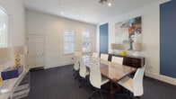 This single-floor meeting space is designed to facilitate professional gatherings in a refined, contemporary setting. At its center sits a distinctive conference table featuring a glass top over a woven rattan base, surrounded by sleek white ergonomic chairs on casters, comfortably accommodating up to eight attendees. The room is finished with crisp white walls and dark charcoal carpeting, creating a clean, polished contrast throughout.

Natural light filters in through multiple windows fitted with white horizontal blinds, offering adjustable brightness to suit any occasion. Recessed ceiling lighting supplements the natural light, ensuring consistent illumination. A bold blue accent wall adds a striking visual element, complemented by a dark wood credenza topped with decorative accessories and a large abstract artwork. A coordinating gallery of four colorful abstract prints adorns another wall, lending the space an artistic, creative energy.

French doors open directly into an adjacent lounge or common area, providing a seamless connection to additional gathering spaces. A secondary door offers private access, enhancing the room's functionality. The overall ambiance strikes a balance between professional utility and contemporary design, making it a versatile and inviting venue for meetings, presentations, or collaborative sessions.