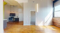 This single-floor unit showcases a compelling mix of industrial heritage and contemporary finishes. The open main living area immediately impresses with warm, original hardwood floors that carry rich character throughout. Soaring ceilings with an arched architectural detail amplify the sense of openness, while abundant natural light streams in, illuminating the bright, neutral walls.

The kitchen is well-appointed with dark wood cabinetry, stainless steel appliances including a gas range, microwave, dishwasher, and refrigerator, complemented by a subway tile backsplash and a modern black-finish faucet. A distinctive structural column adds to the loft-like industrial aesthetic of the kitchen area. Pendant globe lighting on an iron rod fixture adds an artisan touch overhead.

The bedroom offers plush carpeting underfoot and features a standout exposed brick accent wall with large windows that invite natural light and frame exterior views. A closet with a rod and shelf provides practical storage. Soaring ceilings continue here, along with an exposed beam detail that reinforces the building's historic character.

The bathroom is finished with clean white subway tile surrounding a full soaking tub, complemented by hardwood flooring and a simple, modern layout. Throughout the unit, vertical LED lighting fixtures provide a sleek, contemporary accent.