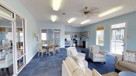 Located at 955 Airport Rd in Destin, Florida, this single-floor community space offers a welcoming and functional environment designed for gathering and relaxation. The open-concept layout seamlessly connects a fully equipped kitchen, a casual dining area, and a comfortable lounge zone, making it well-suited for a variety of social activities.

The kitchen is outfitted with stainless steel appliances, including a refrigerator with ice and water dispenser, a range, and a microwave, complemented by gray cabinetry and ample counter space. Adjacent to the kitchen, the dining area features a round table with cross-back chairs, offering a relaxed setting for shared meals or conversation.

The lounge area is furnished with a neutral-toned sofa and a pair of patterned accent chairs arranged around a small side table, creating an inviting space to unwind. Light blue walls throughout contribute to a calm, coastal atmosphere that reflects the Destin surroundings. Blue modular carpet tiles anchor the seating areas, adding texture and visual interest underfoot.

Natural light filters in through multiple windows dressed with bamboo shades, while flush-mount lighting fixtures and a decorative ceiling fan ensure the space remains bright and comfortable throughout the day. Glass doors offer a visual connection to an adjacent room, enhancing the overall sense of openness.