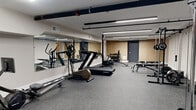 This 820 square foot fitness center, located at 101–209 Court St in New Haven, CT, is a state-of-the-art facility designed for optimal workout experiences. The space features a single floor layout with a thoughtful arrangement of exercise equipment. The room is illuminated by modern LED strip lighting, creating a bright and energizing atmosphere. The floors are covered with high-quality, impact-resistant gray carpet, ideal for various exercise routines. White textured walls contribute to the clean, contemporary aesthetic. The center is equipped with a diverse range of fitness apparatus, including weight machines, free weights, benches, and cardio equipment such as treadmills and stationary bikes. A large mirror spans one wall, allowing users to monitor their form during workouts. The gym also incorporates wooden accent walls, adding warmth to the otherwise sleek design. Security cameras and fire safety equipment are visible, ensuring a safe environment for users. This well-appointed gym provides a comprehensive fitness solution in a compact, efficiently designed space.