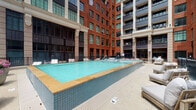 Located in the heart of downtown Columbus, Ohio, this outdoor amenity space at 205 S High St offers a polished urban retreat within a vibrant mixed-use development. The centerpiece of the space is a generously sized rectangular swimming pool finished with teal tile and a warm sandstone coping, lending the area a clean, resort-inspired aesthetic. The pool deck is furnished with contemporary lounge chairs, providing ample space for relaxation. Surrounding the pool on multiple sides is an impressive multi-story building featuring classic red brick facades, large industrial-style windows, and modern balconies—a design that blends historic architectural character with contemporary urban living. The courtyard setting creates a sense of enclosure and privacy while maintaining an open-air atmosphere. The space is defined by its thoughtful contrast of textures: warm brick exteriors, sleek glass, and cool blue water. Accent lighting throughout suggests the space is equally inviting after dark. Situated in Columbus's dynamic downtown core, this amenity area reflects the elevated standard of the surrounding development and offers residents and guests a sophisticated outdoor experience in the middle of the city.