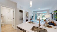 Located at 100 Pine St W in Wilson, NC, this one-bedroom, one-bathroom unit offers a thoughtfully designed open-concept layout that maximizes its efficient footprint. The kitchen is a standout feature, showcasing white shaker cabinetry, a subway tile backsplash, quartz countertops, and a full suite of stainless steel appliances including a refrigerator, range, and over-the-range microwave. A kitchen island with an undermount sink provides additional prep space and flows seamlessly into the bright living area. The living room is bathed in natural light from large windows and a glass door that opens to a private balcony with outdoor views, creating a welcoming and airy atmosphere. Light wood-style flooring runs throughout the main living areas, complementing the neutral, modern aesthetic. The bedroom is a comfortable retreat featuring plush carpet, a ceiling fan, a window with exterior views, and direct access to a dedicated closet. A stacked washer and dryer unit is conveniently tucked within the unit. The bathroom features a white vanity with an undermount sink, a round mirror, and a combination tub and shower. Clean white walls and consistent finishes throughout give the entire space a cohesive, contemporary feel.