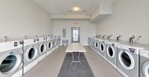 This shared laundry facility is designed for high-capacity, communal use, offering a clean, functional, and well-organized environment. The room is finished with neutral gray walls and smooth flooring, creating a bright and practical atmosphere throughout.

Rows of front-loading washers and dryers line both sides of the room, providing ample machine availability for multiple users simultaneously. Each machine is equipped with individual digital control panels, and utility sinks are conveniently positioned along the same walls for added functionality. A central folding table runs the length of the room, offering generous surface area for sorting and folding laundry. A rolling laundry cart is also available for transporting items with ease.

The space features two entry points: a set of glass double doors that allow natural light to filter in and provide views of an exterior terrace, as well as a secondary gray-framed door with a window. Overhead, flush-mounted and track lighting ensures consistent illumination throughout. A recycling bin and no-smoking signage reflect an emphasis on cleanliness and responsible use. Exit signs and an electrical panel are also present, underscoring the facility's attention to safety and utility.