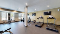 This fitness center is a comprehensively outfitted facility designed to support a full range of workout routines. The space is finished with durable speckled rubber flooring throughout, providing both traction and comfort during exercise. Warm yellow-toned accent walls contrast with crisp white ceilings, creating an energizing yet welcoming atmosphere. Recessed lighting ensures the entire floor plan is consistently and evenly illuminated.

The cardio zone is generously sized, housing multiple treadmills and a rowing machine, alongside elliptical and stationary cycling equipment. Multiple flat-screen televisions are mounted along the walls, allowing users to stay entertained during their workouts. Structural columns are integrated cleanly into the layout without disrupting the flow of the space.

A dedicated strength training area features a cable/pulley machine, a weight bench, and a well-stocked dumbbell rack, with a large wall mirror providing visual feedback for proper form. Additional strength machines are positioned near a bright bank of windows that flood the area with natural light and offer views of the surrounding outdoor environment.

The space is accessible via a wood-paneled door and is thoughtfully organized to allow multiple users to exercise simultaneously without congestion. Overall, the fitness center combines practical functionality with a clean, modern aesthetic.