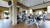 This single-floor fitness center offers a comprehensive workout environment designed to accommodate a range of exercise routines. The space features rich hardwood flooring throughout, lending a warm and polished aesthetic to the room. Warm neutral-toned walls with crown molding details give the gym an elevated, refined appearance beyond a typical workout facility.

The cardio section is well-appointed with multiple stationary bikes, elliptical trainers, and additional aerobic machines arranged to allow comfortable spacing between users. A dedicated free weights area is visible along one wall, complete with a weight rack and bench for strength training exercises. Wall-mounted televisions are positioned for easy viewing during workouts.

Large mirrors line the walls, visually expanding the space and allowing users to monitor their form during exercise. Multiple windows along the walls flood the interior with natural light, creating a bright and energizing atmosphere. Recessed lighting and pendant fixtures supplement the natural light, ensuring the space remains well-lit at all times.

The open layout of the fitness center promotes ease of movement and accessibility to all equipment. Structural columns are integrated thoughtfully into the design, defining zones within the space without disrupting its open feel. Overall, this fitness center combines functional equipment with a sophisticated design.