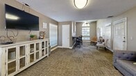 This single-floor space is thoughtfully arranged to accommodate both relaxed lounging and communal dining. Throughout, warm taupe and beige walls are complemented by patterned, earth-toned carpeting and white baseboards, creating a cohesive and welcoming aesthetic. Textured ceilings add a classic touch consistent across all areas.

The living area features a generously sized gray upholstered sofa accented with decorative pillows in chartreuse, navy, and geometric patterns. Wrought iron wall art and framed canvas prints lend personality to the walls, while a rustic wood console table provides functional display space nearby.

An open view leads into a spacious main room, where a wall-mounted television sits atop a white distressed media cabinet. The room flows into a bright sunroom-style nook highlighted by an arched window, where accent chairs and a small side table create an inviting reading or conversation area. Multiple windows dressed with horizontal blinds allow ample natural light throughout.

The dining area accommodates multiple round wooden tables with matching upholstered chairs, suitable for group gatherings. French doors and large windows brighten the space, offering views of the surrounding exterior. The overall ambiance is casual yet polished, with a consistent use of natural wood tones and soft neutral finishes.