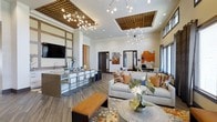 This single-floor space showcases a thoughtfully designed open-concept layout that combines a comfortable lounge area with a functional kitchen bar. The soaring ceilings draw the eye upward to a pair of striking sputnik-style chandeliers and recessed wood-panel ceiling insets with integrated lighting, adding architectural depth and warmth to the space. The expansive layout is anchored by a large, light-toned sectional sofa adorned with earth-toned accent pillows in burnt orange and warm brown, complemented by a bold black-and-white circular area rug beneath a decorative floral centerpiece. Adjacent to the lounge, a sleek kitchen bar features a stone-topped island with modern bar seating, a wall-mounted television, and a sink, making it ideal for both casual gatherings and events. Wide-plank wood-look flooring flows throughout, tying the zones together cohesively. Artwork in rich amber and blue hues punctuates the white walls, reinforcing the warm, contemporary color palette. Natural light filters through windows along the perimeter, enhancing the bright and welcoming ambiance of this well-appointed communal space.