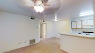This single-floor residential space presents a straightforward and functional layout designed for comfortable everyday living. The main living area is carpeted throughout and features neutral white walls that create a bright, clean atmosphere. A ceiling fan with an integrated light fixture is centrally mounted, providing both airflow and ambient lighting to the space.

A partial-wall breakfast bar separates the living area from the adjacent kitchen, offering a convenient pass-through that maintains an open feel between the two spaces. The kitchen area includes what appears to be a framed mirror or cabinet panel, adding a modest decorative detail to the room.

Multiple HVAC vents are positioned across the floor and ceiling, ensuring efficient climate control throughout the unit. A doorway leads to a hallway that connects to additional rooms, while a secondary entrance door is flanked by a window fitted with horizontal blinds, allowing natural light to filter in while maintaining privacy. The window provides a glimpse of greenery outside, adding a subtle connection to the outdoors.

Overall, the space offers a clean, move-in-ready environment with a neutral palette that serves as a blank canvas for personalization. The practical layout, consistent flooring, and thoughtful ventilation make it a functional and adaptable living space.