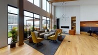 Located at 625 Fourth Ave S in Minneapolis, this single-floor space delivers an impressive blend of modern design and functional amenities. The layout is open and expansive, anchored by soaring double-height ceilings that flood the interior with natural light and create an airy, sophisticated atmosphere. Warm natural wood flooring runs throughout, complementing exposed timber columns and wood-trimmed architectural details that add warmth to the otherwise contemporary aesthetic.

The lounge and gathering area is a standout feature, furnished with comfortable seating arrangements and accented by a sleek linear fireplace set into a built-in niche. A geometric pendant light fixture adds a sculptural element overhead. Floor-to-ceiling black-framed windows along one wall open the space visually to a landscaped outdoor terrace, offering panoramic views of the surrounding Minneapolis skyline.

The fully equipped kitchen area is thoughtfully designed with gray cabinetry, a striking blue-toned tile backsplash framed in warm wood, white quartz countertops, and full-size appliances including a refrigerator, microwave, and dishwasher. A large central island with seating provides additional functionality. Curated artwork displayed throughout the space adds a gallery-like quality to the overall ambiance.
