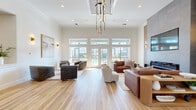 This thoughtfully designed single-floor space combines modern elegance with an open, airy atmosphere. The expansive living room serves as the centerpiece, boasting soaring ceilings adorned with crown molding and a striking tiered chandelier that anchors the room. Wide-plank light hardwood flooring flows throughout, enhancing the sense of openness and warmth.

Floor-to-ceiling windows and a set of French doors flood the living area with abundant natural light while offering direct views of an outdoor pool area, creating a seamless indoor-outdoor connection. The living room is anchored by a full-height stone tile accent wall featuring a sleek linear fireplace and a mounted flat-screen television, flanked by vertical slatted wood paneling for added texture and depth. Multiple seating arrangements — including leather sofas, swivel accent chairs, and a round side table — make the space both functional and inviting.

A dedicated lounge nook near the French doors provides an intimate setting for conversation, complete with wall sconces that lend a warm, ambient glow and curated wall art that adds a refined aesthetic touch.

The kitchen is clean and efficient, featuring white cabinetry, light stone countertops, open natural wood floating shelves, and stainless steel appliances — a minimalist design that prioritizes both form and function.