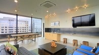 This contemporary single-floor space showcases a thoughtfully designed open-plan layout that seamlessly blends functionality with modern aesthetics. The interior is anchored by a generous lounge area featuring a sleek, low-profile coffee table, comfortable seating, and a wall-mounted flat-screen television, creating an ideal setting for both relaxation and presentations.

A built-in kitchenette lines one wall, fitted with light wood cabinetry, a sink, and a dark backsplash panel that adds a refined contrast to the bright, neutral palette throughout. Adjacent to the kitchenette, a dedicated dining area accommodates a full table with multiple chairs, making it well-suited for group gatherings or working sessions.

Floor-to-ceiling windows along one side of the space flood the interior with natural light while offering expansive views of the surrounding urban skyline, lending the room an elevated, city-connected atmosphere. Recessed lighting and a ceiling-mounted HVAC unit ensure year-round comfort, while track lighting adds a layer of adjustable ambiance. The polished concrete-style flooring and clean white walls contribute to the space's minimalist, professional character, making it a versatile environment suitable for a variety of uses.