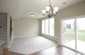 This two-story home presents a thoughtfully designed main floor where the living, dining, and kitchen areas flow together in an open-concept layout. The living area features plush carpeting and warm neutral-toned walls, creating a welcoming atmosphere. Natural light pours in through a large multi-pane window and a glass exterior door that opens to the backyard, offering pleasant outdoor views of a green lawn. An elegant chandelier with dark metal detailing serves as a focal point in the dining area, complementing the rich dark hardwood flooring that defines the kitchen and dining space.

The kitchen is equipped with dark cabinetry, stainless steel appliances including a refrigerator and microwave, and a central island with pendant lighting overhead. A staircase with dark metal balusters and carpeted treads leads to the second floor, positioned adjacent to the kitchen for convenient access. Flush-mount ceiling fixtures provide additional lighting throughout the main level. The overall design balances functionality with a clean, modern aesthetic, making excellent use of natural light and cohesive finishes throughout.