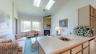 This single-floor residence offers a thoughtfully designed layout that combines comfort and functionality throughout its well-appointed spaces. The living room is a standout feature, boasting soaring vaulted ceilings, dual skylights, and a ceiling fan that fill the room with natural light and a sense of openness. Large windows frame pleasant outdoor views of lush greenery, while a tiled fireplace serves as a warm focal point. Soft teal carpeting runs throughout much of the home, lending a cohesive and inviting atmosphere.

The dining area is elegantly defined by a chandelier overhead and connects seamlessly to the kitchen, which features white cabinetry with wood trim, ample counter space, a range, dishwasher, and a pass-through window that adds charm and connectivity to the adjoining spaces. A second sitting or den area offers additional flexible living space with vaulted ceilings and a bright window.

The bedrooms are generously sized with plush carpeting and direct access to the bathrooms. The full bathroom includes a wide vanity with ample storage and a sliding glass shower enclosure. A separate half bathroom with patterned tile flooring provides added convenience. The overall ambiance is bright, airy, and welcoming throughout.