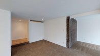 This single-floor space presents a straightforward, functional layout with several notable architectural details. The main living area is finished with wall-to-wall neutral-toned carpeting, contributing a warm, understated feel underfoot. White walls throughout keep the interior bright and versatile, providing a blank canvas adaptable to various design preferences.

A prominent dark brick fireplace column serves as a defining architectural feature, adding texture and a sense of character to the otherwise clean-lined space. Its placement creates a natural visual anchor and subtly divides the room, offering an organic sense of spatial definition without fully closing off the floor plan.

The ceiling is smooth and white, with recessed lighting integrated to provide even, functional illumination throughout the room. A stepped platform or raised area is visible along one wall, hinting at an intentional variation in floor levels that adds subtle architectural interest and potential for differentiated use zones within the space.

The overall ambiance is neutral and move-in ready, with a simple, practical design that lends itself well to both residential and flexible-use arrangements. The absence of ornate detailing keeps the focus on structural elements like the brick column and the layered floor levels.