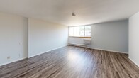 This single-floor unit offers a straightforward, functional layout finished with consistent wood-look laminate flooring throughout. The main living area is open and bright, featuring a large window with a baseboard heating unit beneath it, allowing natural light to fill the space. Crisp white walls and clean white baseboards contribute to a neutral, move-in-ready aesthetic.

A dedicated bedroom provides generous floor space and includes a built-in double closet with louvered bifold doors, offering practical storage. The room maintains the same neutral color palette and laminate flooring seen throughout the unit.

The bathroom is efficiently designed, featuring a white vanity with an integrated sink and cabinet storage below, a wall-mounted mirror with overhead lighting, and a toilet. Wood-look flooring continues into this space, tying the unit together cohesively. A louvered bifold door and a floor vent are also present, adding to the room's practicality.

Overall, the unit presents a clean, minimal aesthetic with consistent finishes across all spaces. The neutral tones, functional closet storage, and natural light from the living area window make it a versatile and practical space.