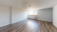 This single-floor unit offers a straightforward, functional layout finished with consistent wood-look laminate flooring throughout. The main living area is open and bright, featuring a large window with a baseboard heating unit beneath it, allowing natural light to fill the space. Crisp white walls and clean white baseboards contribute to a neutral, move-in-ready aesthetic.

A dedicated bedroom provides generous floor space and includes a built-in double closet with louvered bifold doors, offering practical storage. The room maintains the same neutral color palette and laminate flooring seen throughout the unit.

The bathroom is efficiently designed, featuring a white vanity with an integrated sink and cabinet storage below, a wall-mounted mirror with overhead lighting, and a toilet. Wood-look flooring continues into this space, tying the unit together cohesively. A louvered bifold door and a floor vent are also present, adding to the room's practicality.

Overall, the unit presents a clean, minimal aesthetic with consistent finishes across all spaces. The neutral tones, functional closet storage, and natural light from the living area window make it a versatile and practical space.