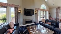 The Bound Brook Club Room at 675 Tea Street is a well-appointed, single-floor community space designed for socializing and recreation. The room's most striking architectural feature is its soaring vaulted ceiling, which creates an open, airy atmosphere and is complemented by large windows that flood the interior with natural light and offer views of the surrounding green landscape.

The main lounge area is anchored by a sleek linear fireplace topped with a wall-mounted flat-screen television, flanked by wall sconces and dressed with decorative patterned drapery. Plush leather seating and a star-patterned area rug contribute to the room's classic Americana aesthetic, further accented by displayed flags and themed décor.

A dedicated game area features a billiards table, providing additional recreational options for residents. Adjacent to the lounge, a well-equipped kitchenette serves as a central hub for gatherings, featuring rich dark wood cabinetry with glass-front upper doors, granite countertops, pendant lighting, and appliances including a coffee station and microwave. A peninsula counter with bar seating in bold red adds a social focal point.

A separate music nook houses a grand piano positioned beside large picture windows overlooking open green space, ideal for quiet enjoyment or entertainment. Hardwood-style flooring flows throughout, tying the various zones together into a cohesive and inviting community retreat.