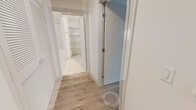 This contemporary single-floor residence is thoughtfully designed with a clean, modern aesthetic throughout. Light wood-tone flooring flows seamlessly across all areas, complemented by crisp white walls that create a bright and airy atmosphere.

The kitchen is a standout feature, equipped with full stainless steel appliances including a refrigerator, range, microwave, and dishwasher. Light wood cabinetry extends floor to ceiling, offering generous storage, while white quartz countertops and a white subway tile backsplash add a polished finish. A kitchen island provides additional prep and dining space.

The living and bedroom areas are generously sized, each benefiting from large windows dressed with black roller shades that admit abundant natural light and offer views of the surrounding modern building complex. Select rooms provide direct access to an outdoor balcony area through glass-panel doors.

The bathroom is finished to a high standard, featuring a floating vanity with light wood cabinetry, a large mirror, open shelving, and a tiled tub-shower combination. A dedicated walk-in closet includes built-in shelving and hanging rods for well-organized storage. A corridor with additional open shelving connects the living spaces efficiently, completing this well-planned and stylish residence.