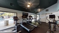 Located at 2105 Ravenglass Pl in Raleigh, NC, this single-floor fitness center is a well-equipped workout facility designed to support a full range of exercise routines. The space features a striking dark teal ceiling with ceiling fans and recessed lighting, creating a focused and energizing atmosphere complemented by white walls and durable rubber flooring throughout.

The cardio area is outfitted with multiple treadmills, an elliptical trainer, a stationary bike, and a stair climber, all positioned to take advantage of the natural light streaming in through several windows with outdoor views. A wall-mounted flat-screen television is centrally positioned for entertainment during workouts.

The strength training section includes a multi-function cable machine, a weight bench, and a well-stocked dumbbell rack offering a range of weights to accommodate various fitness levels. A sliding glass barn-style door leads to an adjacent studio space, which features warm hardwood flooring, frosted glass partitions with decorative striping, and high transom windows that flood the area with natural light — ideal for stretching, yoga, or group exercise.

The overall layout is efficient and thoughtfully organized, providing a comprehensive fitness experience within a compact, purpose-built space.