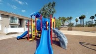 This outdoor community space is defined by its lively, well-equipped playground area and open grounds, situated within a residential complex characterized by stucco-exterior buildings. The playground is the centerpiece of the space, showcasing a large, multi-component play structure in bold blue, red, and yellow. It features multiple slides — including a wide blue slide and a curved grey slide — along with climbing elements, tunnels, and interactive panels, all set atop a rubberized safety surface that provides cushioning underfoot. The surrounding landscape is dotted with mature palm trees and leafy shade trees, adding a natural canopy against the clear blue sky. White vinyl privacy fencing defines boundaries within the property, creating a sense of separation between activity zones. Exterior staircases with metal railings lead to upper-level units of the multi-story stucco buildings, which feature sliding glass doors and textured beige facades. Open paved walkways connect the various areas of the complex. The broader grounds offer views of open lots and neighboring low-rise structures, consistent with a warm, arid climate setting. Overall, the space presents a functional and community-oriented outdoor environment with a strong emphasis on recreational amenities.