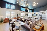 Located at 1551 Riverside Pkwy in Douglasville, GA, this single-floor community lounge space offers an open-concept layout that seamlessly combines a kitchen and living area into one cohesive, inviting environment. The space is defined by its dramatically high ceilings finished in a deep navy-gray tone, which contrast beautifully against crisp white board-and-batten walling below, creating a sophisticated two-tone architectural aesthetic. Rows of clerestory windows line the upper walls, flooding the interior with abundant natural light and offering glimpses of the surrounding neighborhood. Statement sculptural chandeliers with exposed Edison-style bulbs serve as striking focal points throughout the living area, complementing the warm honey-toned hardwood flooring below. The living zone features multiple comfortable seating arrangements, including plush sectional sofas and leather accent chairs, alongside marble-topped bistro tables ideal for social gatherings. The kitchen is a standout feature, anchored by a generously sized white waterfall island with seating for several guests outfitted with black and gold barstools. Dark wood cabinetry, subway tile backsplash, open industrial-style shelving, and pendant lighting complete the kitchen's modern yet warm character. Glass-paned interior doors connect to adjacent spaces, enhancing the open, airy flow throughout.