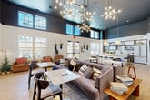 Located at 1551 Riverside Pkwy in Douglasville, GA, this single-floor community lounge space offers an open-concept layout that seamlessly combines a kitchen and living area into one cohesive, inviting environment. The space is defined by its dramatically high ceilings finished in a deep navy-gray tone, which contrast beautifully against crisp white board-and-batten walling below, creating a sophisticated two-tone architectural aesthetic. Rows of clerestory windows line the upper walls, flooding the interior with abundant natural light and offering glimpses of the surrounding neighborhood. Statement sculptural chandeliers with exposed Edison-style bulbs serve as striking focal points throughout the living area, complementing the warm honey-toned hardwood flooring below. The living zone features multiple comfortable seating arrangements, including plush sectional sofas and leather accent chairs, alongside marble-topped bistro tables ideal for social gatherings. The kitchen is a standout feature, anchored by a generously sized white waterfall island with seating for several guests outfitted with black and gold barstools. Dark wood cabinetry, subway tile backsplash, open industrial-style shelving, and pendant lighting complete the kitchen's modern yet warm character. Glass-paned interior doors connect to adjacent spaces, enhancing the open, airy flow throughout.
