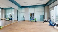 This fitness studio is designed with both functionality and aesthetics in mind, offering a bright, open environment well-suited for a variety of exercise activities. The space features light hardwood flooring throughout, providing a clean and resilient surface ideal for stretching, aerobics, yoga, or general training. Tall, frosted glass partition walls line the perimeter, flooding the interior with diffused natural light while maintaining privacy and creating a sense of spaciousness.

A bold teal accent band runs along the upper portion of the white walls, adding a vibrant pop of color that energizes the space and gives it a polished, contemporary look. The ceiling is finished in white and incorporates recessed lighting and ceiling-mounted speakers, ensuring both ample illumination and an acoustically comfortable environment.

A fitness accessory rack holds a collection of medicine balls in varying weights, a stability ball, and additional workout tools, making the space practical and organized. A stationary bike positioned near the glass wall rounds out the available equipment. A wall-mounted mirror on one side allows users to monitor their form during exercise. Overall, the studio conveys a clean, professional, and motivating atmosphere.