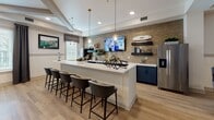 Located at 161 Grande Oaks Drive in Charleston, South Carolina, this single-floor community clubhouse offers a well-appointed gathering space designed for comfort and socialization. The interior showcases warm light hardwood flooring that flows seamlessly throughout, tying together the various distinct areas of the space.

A welcoming lounge area features a sleek wall-mounted electric fireplace set against a bold slate-blue accent wall, accompanied by a mounted flat-screen TV, a tufted leather bench, and a graphic black-and-white area rug. Decorative framed artwork adorns the surrounding walls, adding personality and visual interest.

The kitchen and bar area serves as a central hub, anchored by a generous white quartz island with bar seating for multiple guests. Striking teal cabinetry lines the walls beneath open dark shelving, all set against a warm exposed brick accent wall. Pendant lighting with glass shades illuminates the space, which also includes a stainless steel refrigerator and a dedicated coffee and refreshment station — ideal for casual gatherings.

Adjacent to the kitchen, an elegant lounge area features plush, dark velvet wingback chairs and sofas arranged around a round white coffee table. Large windows with Roman shades and draping dark curtains invite natural light while a textured mosaic accent wall and cozy banquette seating provide a refined, welcoming ambiance.