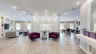The Residents Lounge at 20 Windsor Rd in Belle Mead, NJ is a well-appointed communal gathering space designed to foster comfort and community. The single-floor layout is unified by light wood-tone herringbone-patterned flooring that flows seamlessly throughout, creating a cohesive and inviting atmosphere.

A striking decorative lattice partition with an Art Deco-inspired fan motif serves as a central architectural feature, elegantly dividing the open floor plan into distinct functional zones without fully closing off the space. Natural light filters in through multiple windows dressed with neutral Roman shades, brightening the interior alongside recessed ceiling lighting.

The lounge area features plush burgundy armchairs and a sofa seating arrangement, complemented by a built-in shelving unit housing a flat-screen television, books, and curated décor — ideal for relaxed gatherings. A separate social seating nook with softer furnishings offers a quieter retreat.

The kitchen and dining area includes a long counter-height bar with purple upholstered stools, pendant lighting, open display shelving, and a mounted television, alongside adjacent dining tables for more formal socializing. Colorful artwork punctuates the walls throughout, adding vibrancy to the neutral palette. A glass double-door entry provides a welcoming arrival experience, completing this thoughtfully designed residents' amenity space.